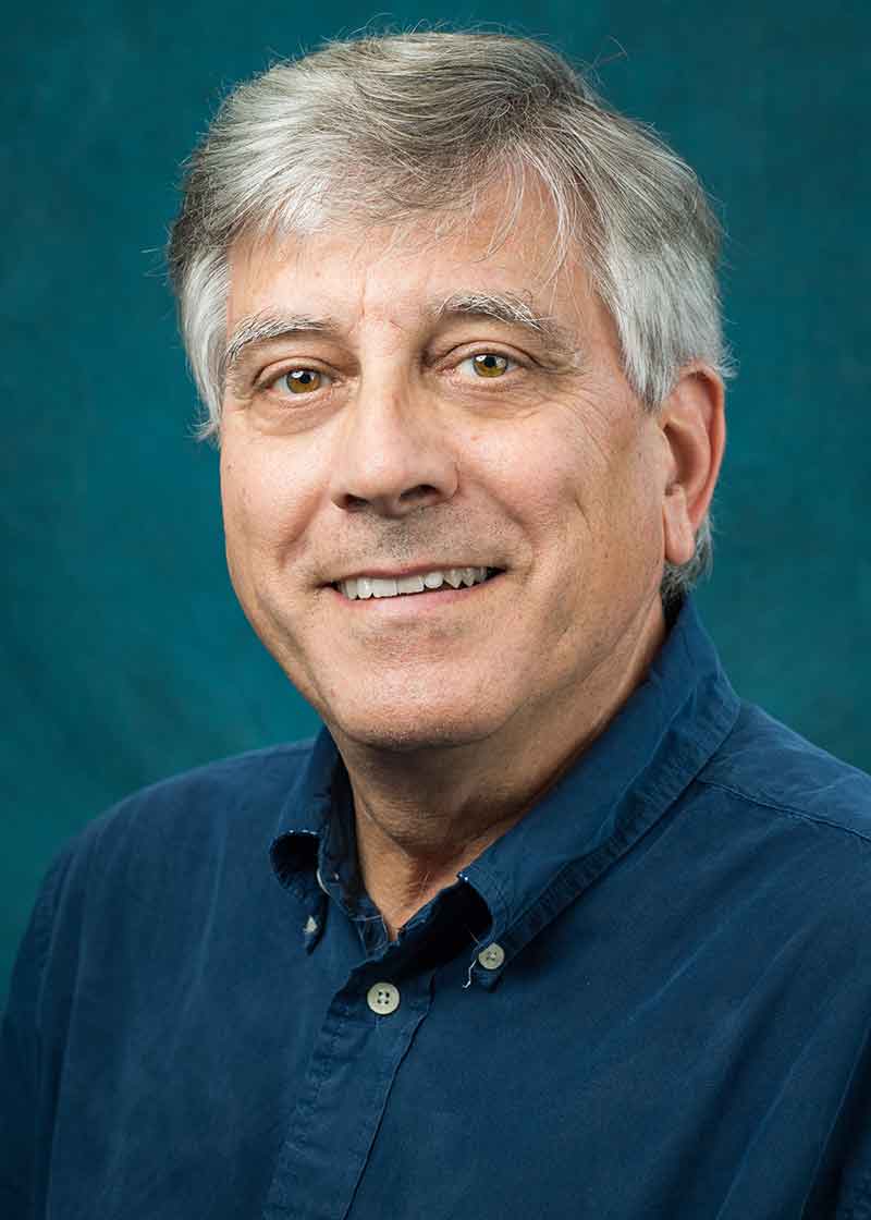 A studio portrait of a grey-haired man in a blue-green button-down shirt.