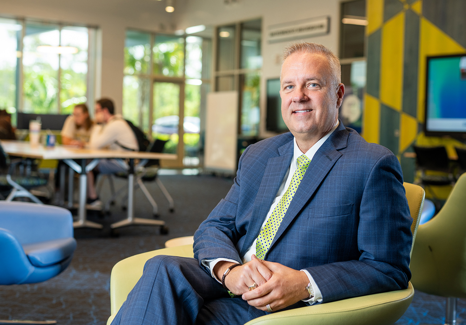 Man in blue suit and green tie sitting in a chair in FGCU's Lucas Hall.