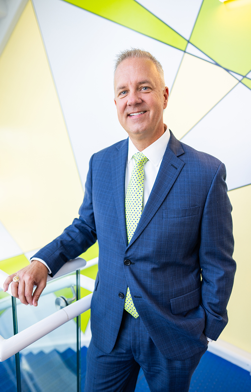 Man in blue suit and green tie standing against a geometric background.