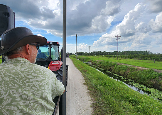 A man in a cowboy hat and green shirt, seated behind a big red tractor, looks our at a green farm as the clouds roll overhead.