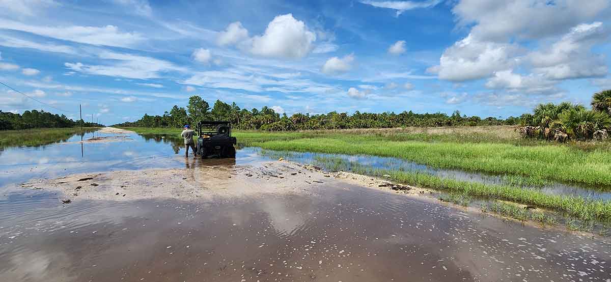 A man in a blue cap stands on the driver's side of an ATV, looking off into the distance, on a dirt road half under water. The right side of the photo features a grassy and forested area and the top half of the picture is blue sky with strings of puffy, white clouds. On the left, power lines are visible but partially submerged in water.