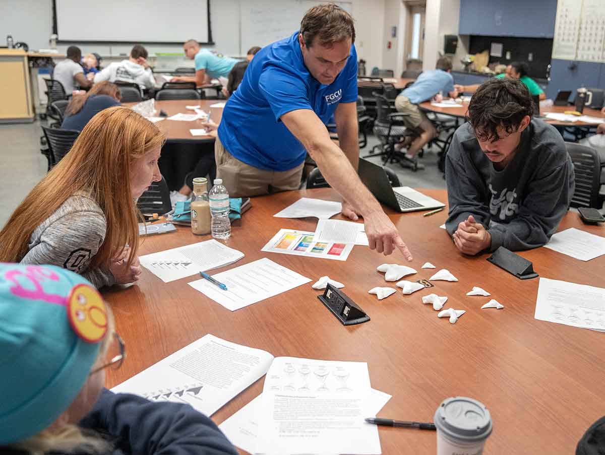 A man in a blue polo stands, pointing at white 3D-printed teeth on a large table, with students to either side