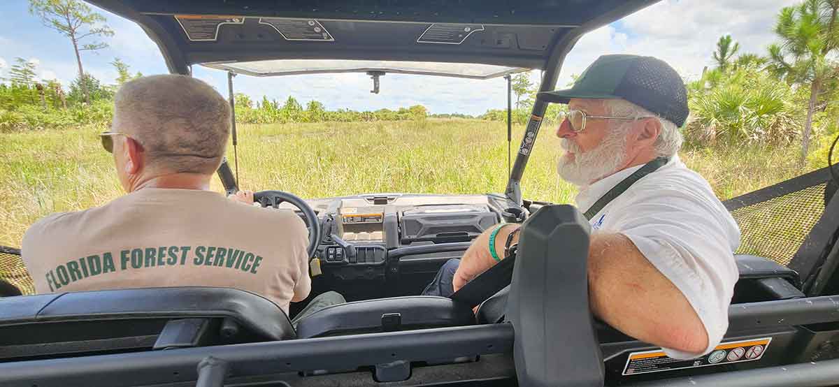 A white-bearded man in a baseball cap and white button-down shirt sits in the passenger seat of a covered ATV looking toward the driver, who is in Ray-Ban style sunglasses and a beige t-shirt that reads Florida Forest Service. In front of the ATV is a field of grass and trees. View is from behind the two men, from the back seat.