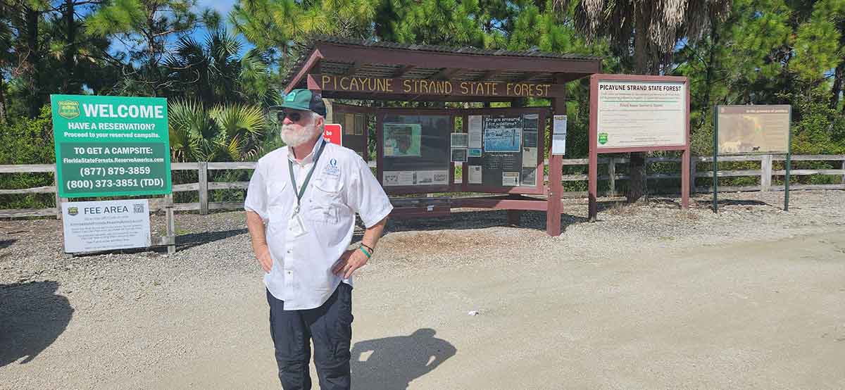 A man with a white beard in a hat and white button-down shirt stands in front of a sign that reads Picayune Strand State Forest