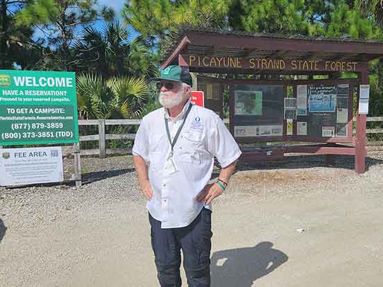 A man with a white beard in a hat and white button-down shirt stands in front of a sign that reads Picayune Strand State Forest