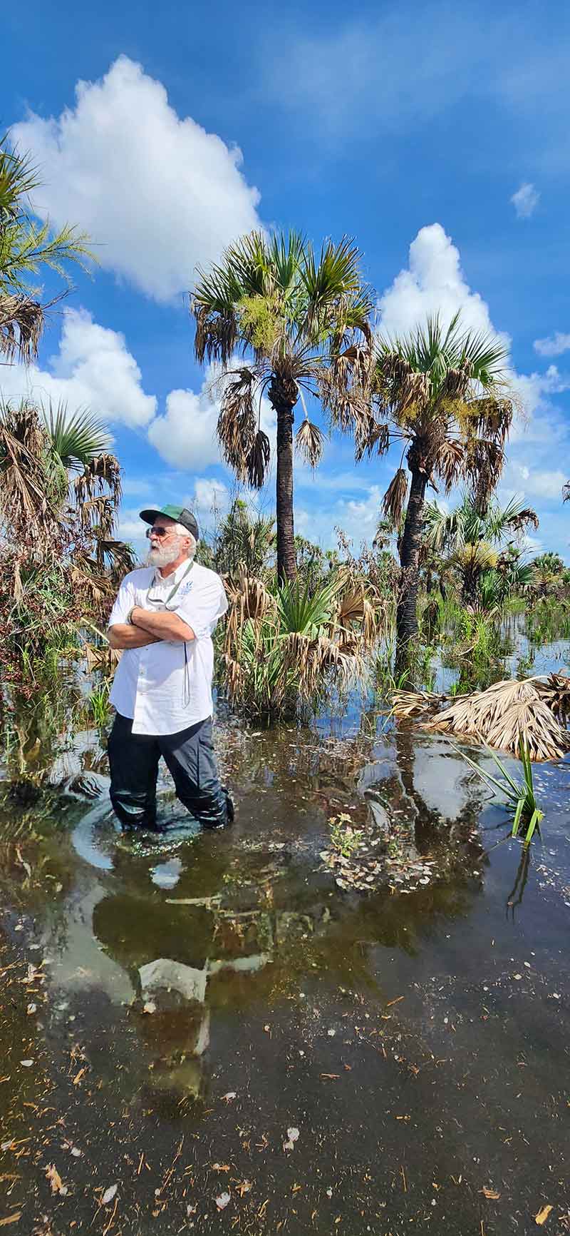 A white bearded man in a green baseball cap stands with his arms folded over his chest in knee-deep water, staring off camera to his right