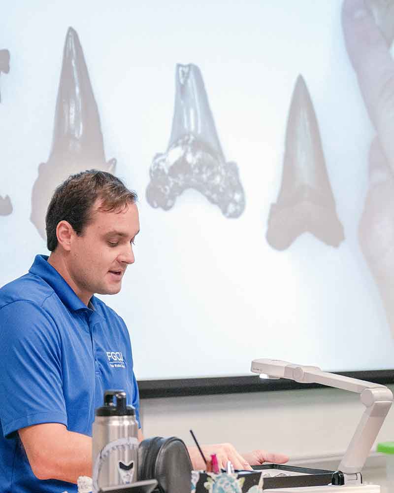 A man in a blue polo stands at a podium with a projected image behind him of megalodon teeth