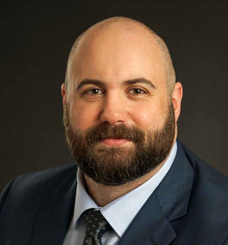 A professional headshot of a bearded man wearing a white button-down shirt, tie and a black blazer