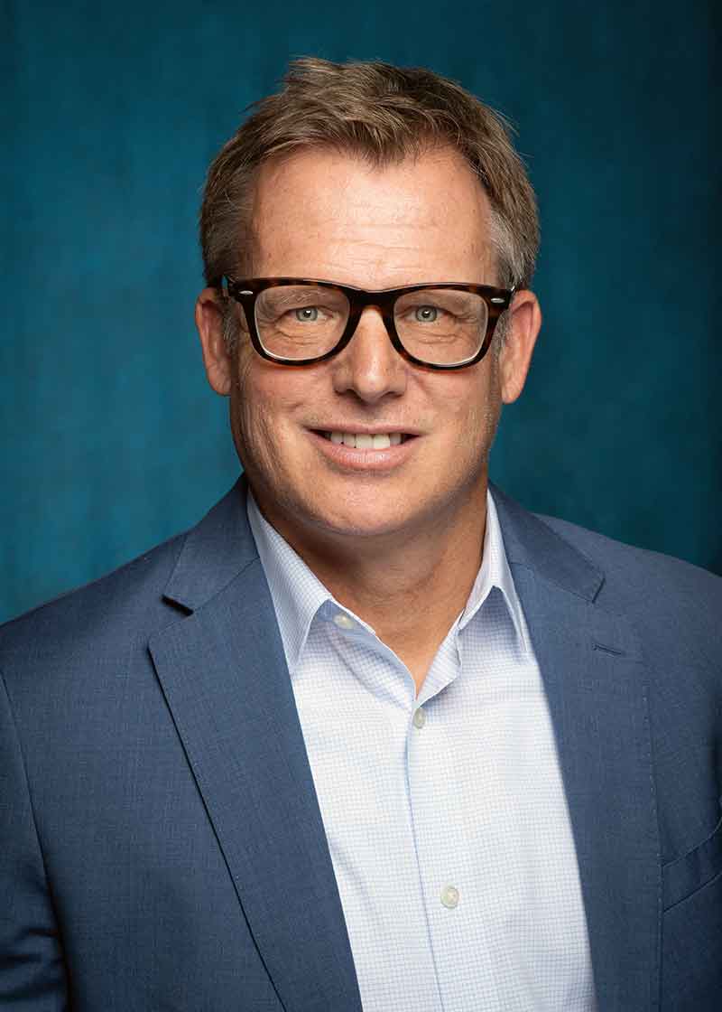 A professional headshot of a man in glasses, wearing a pale blue button-down shirt and a grey-blue blazer