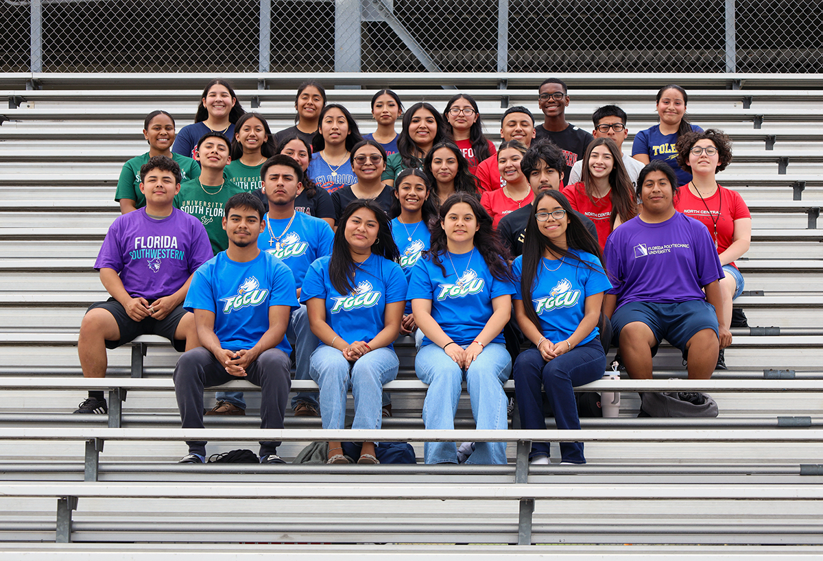 A group of high school students in college T-shirts sit on bleachers