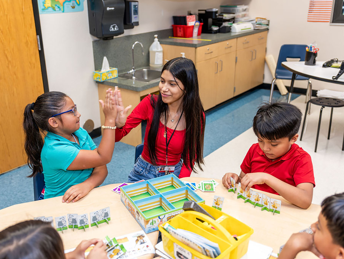 A woman sits at a desk in a classroom with two younger children