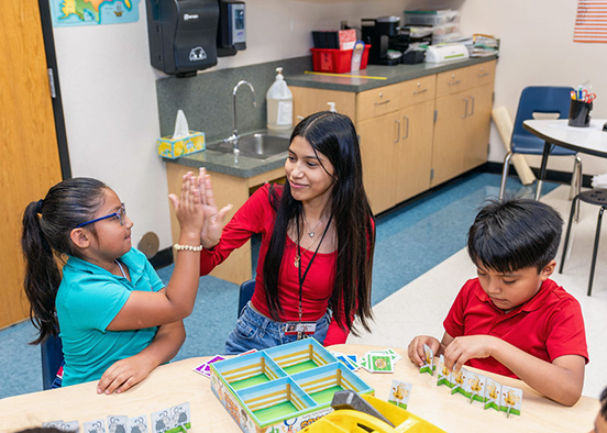 A woman sits at a desk in a classroom with two younger children