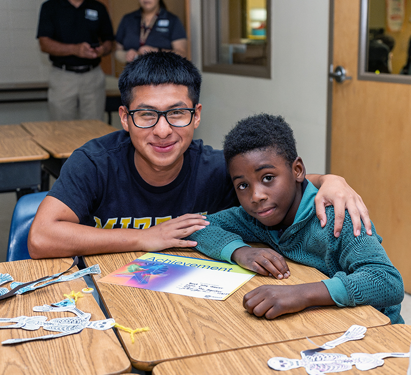 A young man and a boy sit at a desk in a classroom