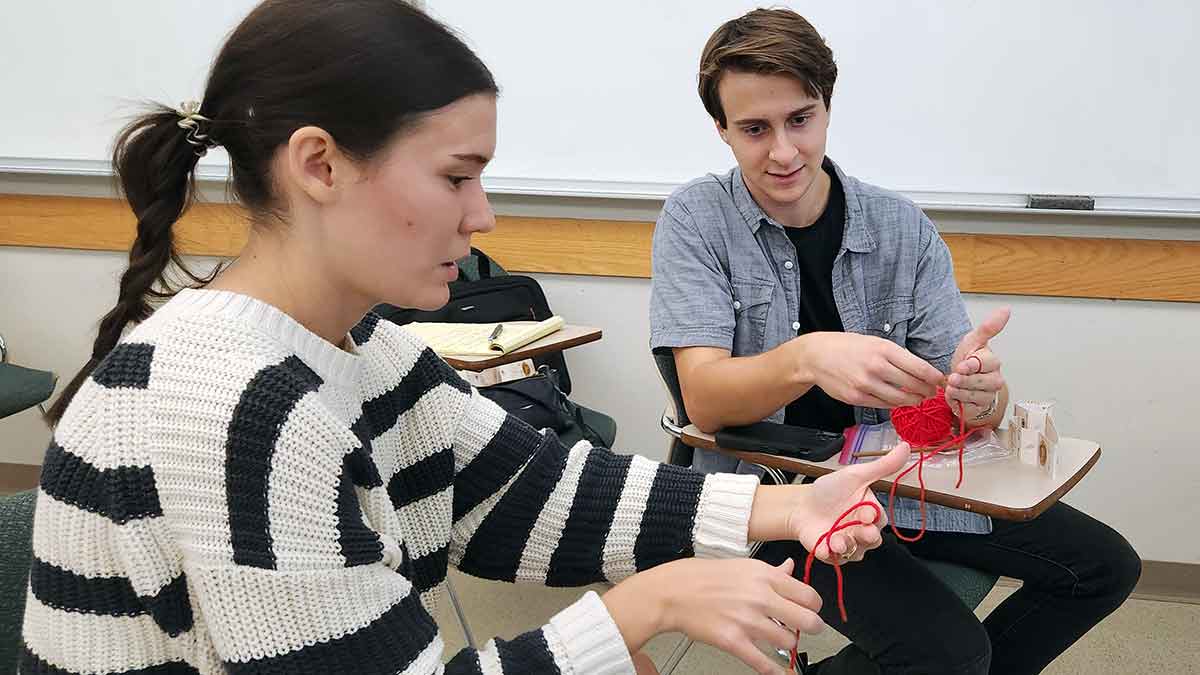 A woman in a white and black sweater and a man in a grey button-down shirt over a black t-shirt sit in desk-chairs in a classroom, with red yarn and crochet needles in their hands