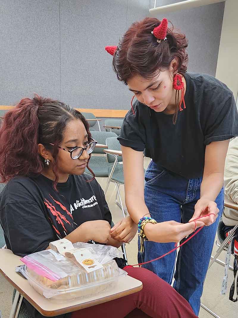 Two women in black t-shirts in a classroom, one standing and demonstrating a knitting technique with red yarn to one seated