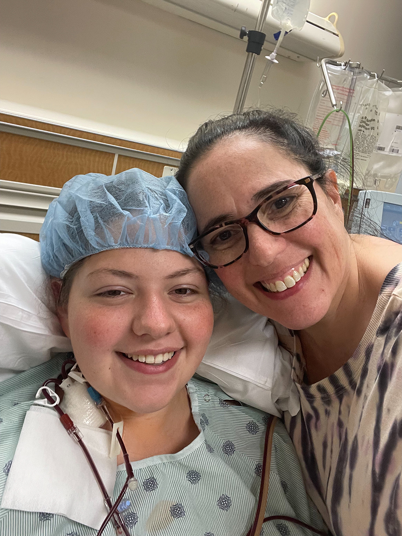 A young, smiling woman in a hospital bed wears a protective cap while she and her smiling mom pose for a selfie