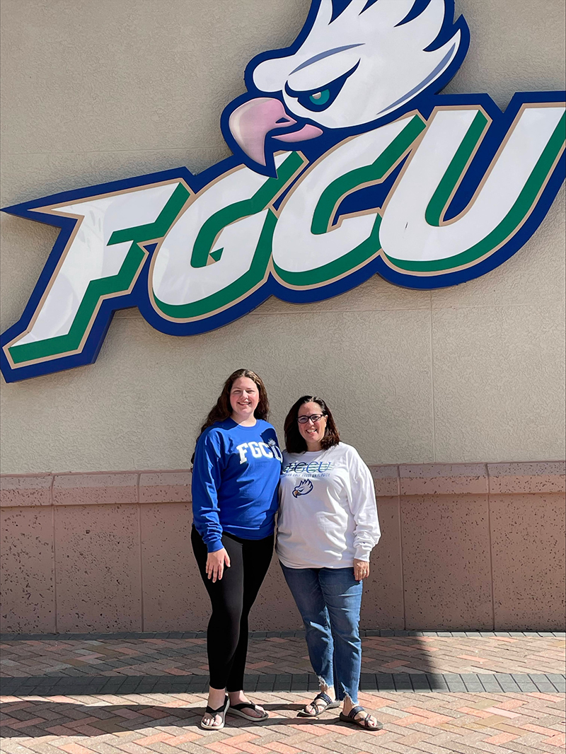 Below a large FGCU logo on an outdoor wall, A young woman in a blue FGCU sweatshirt and her mom, wearing a white FGCU long-sleeve shirt, pose for the camera