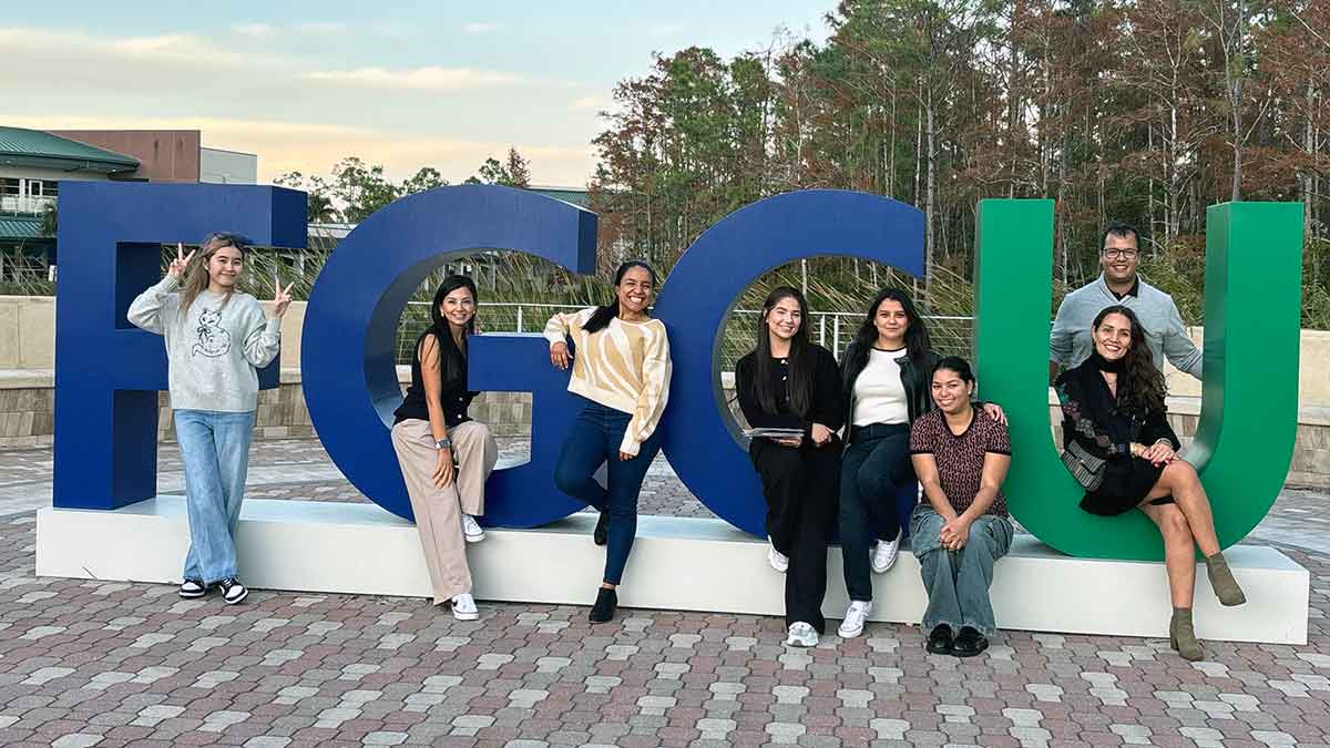 Eight students stand or sit outdoors in the giant, full color university logo with FGC in blue and U in green