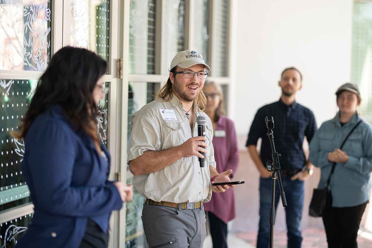 A man with medium-length hair and a beard wears glasses, a baseball cap, a beige button-up shirt and khakis, holds a microphone and is flanked by three people in front of a window with visible white dots