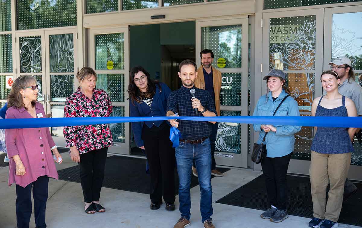 A man in jeans and a blue button-up shirt holds a microphone in his left hand and cuts a blue ribbon with scissors in his right hand in front of windows featuring a white design of dots and a nature scene as six people look on