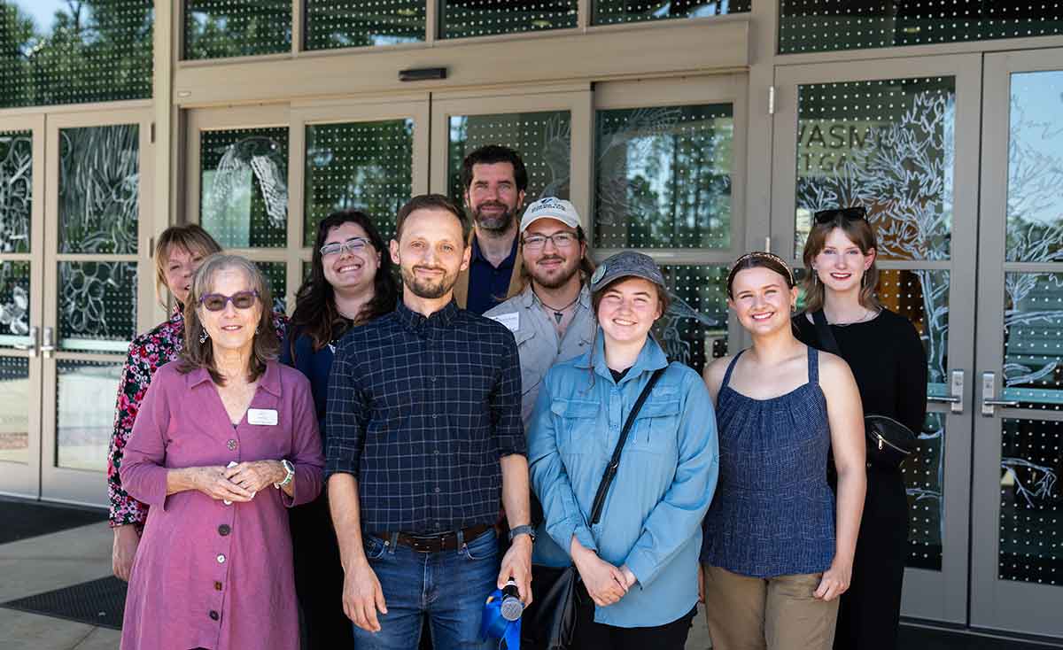 A group of nine people pose in front of an artistic window design of birds and trees and a grid of dots