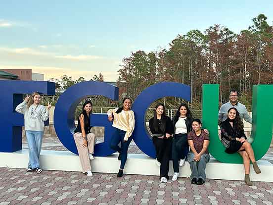 Eight students stand or sit outdoors in the giant, full color university logo with FGC in blue and U in green