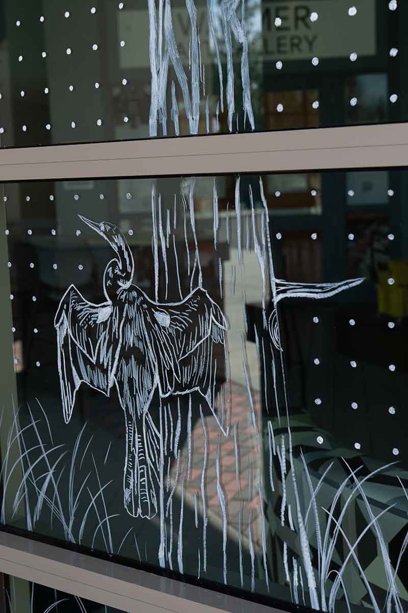 A close-up of the design on a window is of a line drawing of tree bark and an anhinga bird spreading its wings with grass shooting up around it, surrounded by a grid of white dots