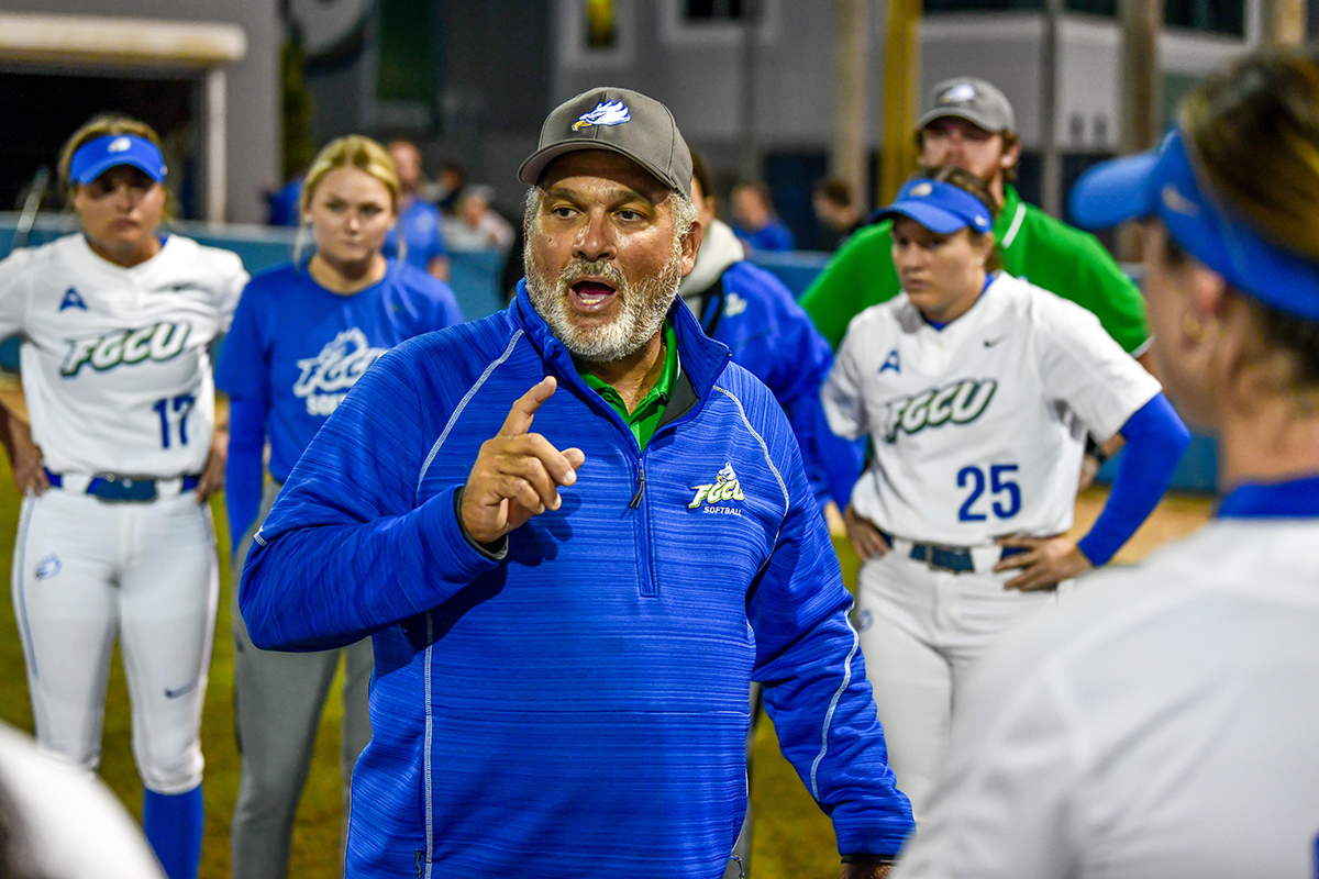 Male coach in blue top talking to female athletes in uniform on ballfield