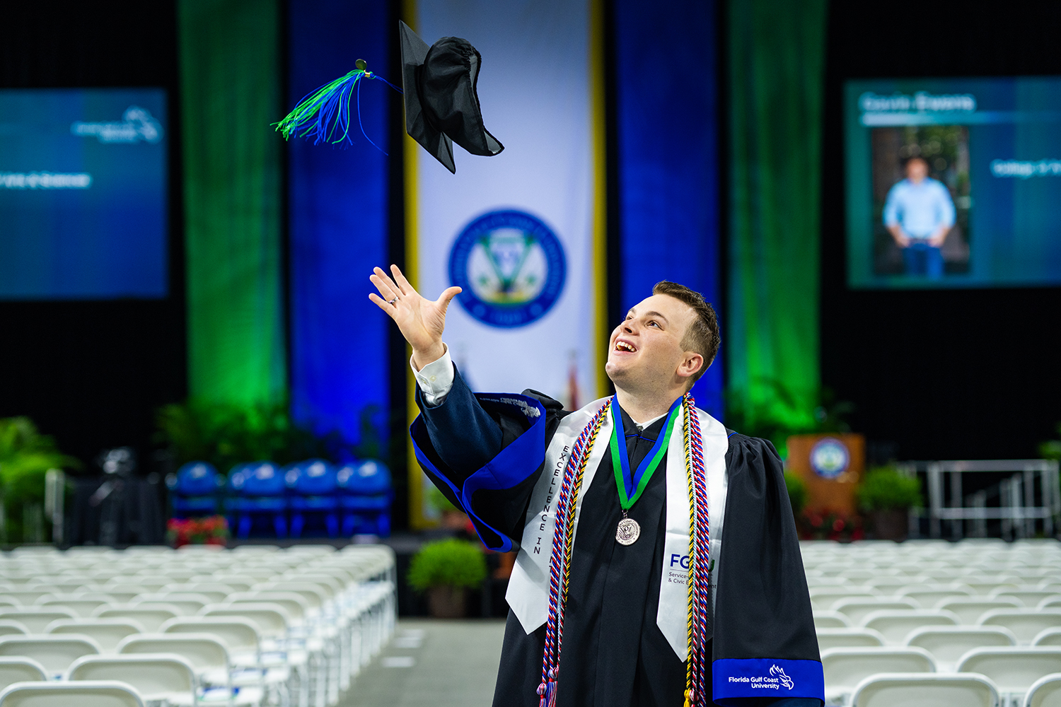 Man in commencement robe and sashes throwing his mortarboard in the air