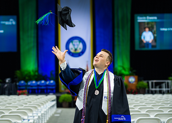 Man in commencement robe and sashes throwing his mortarboard in the air