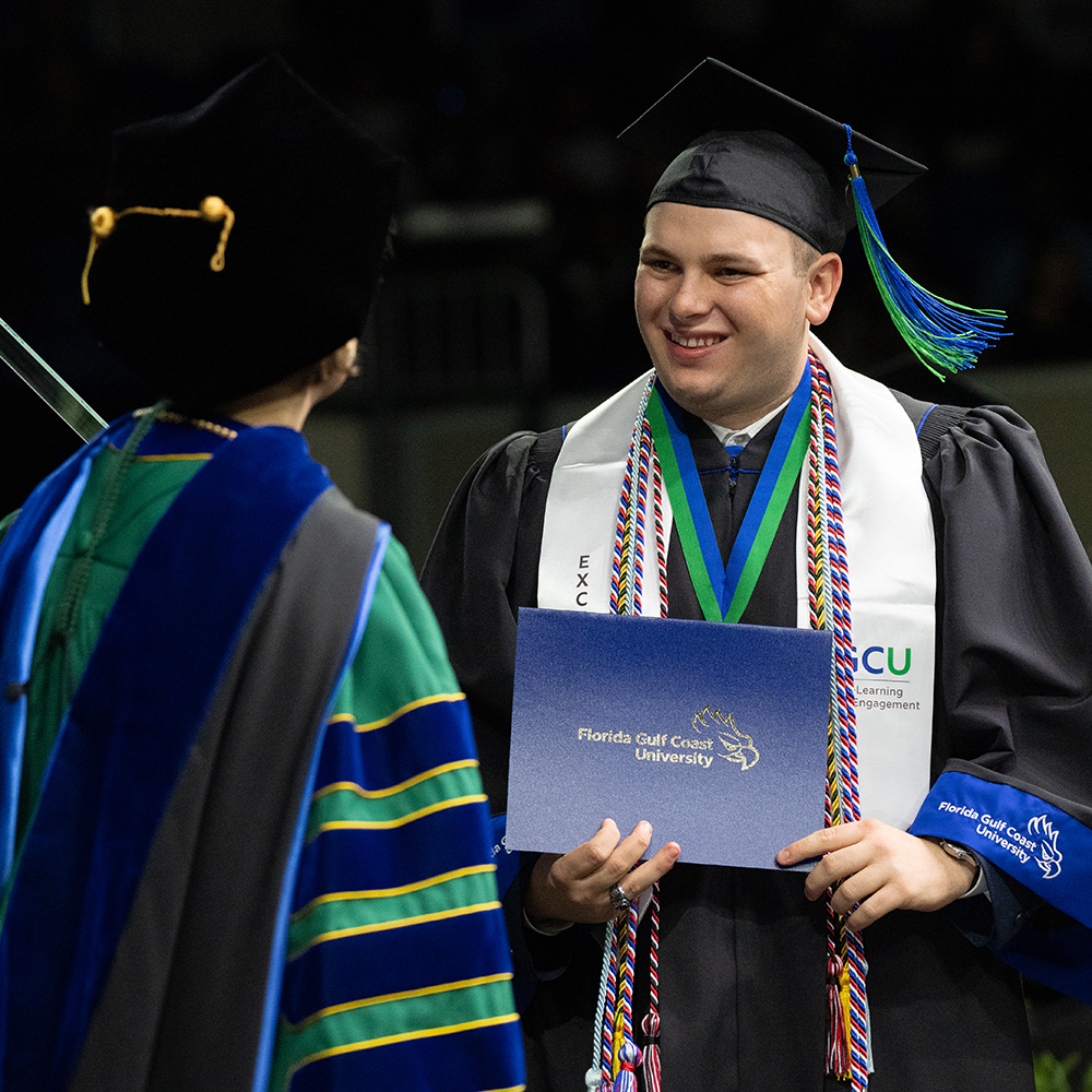 Man in commencement robe and sashes receiving his diploma