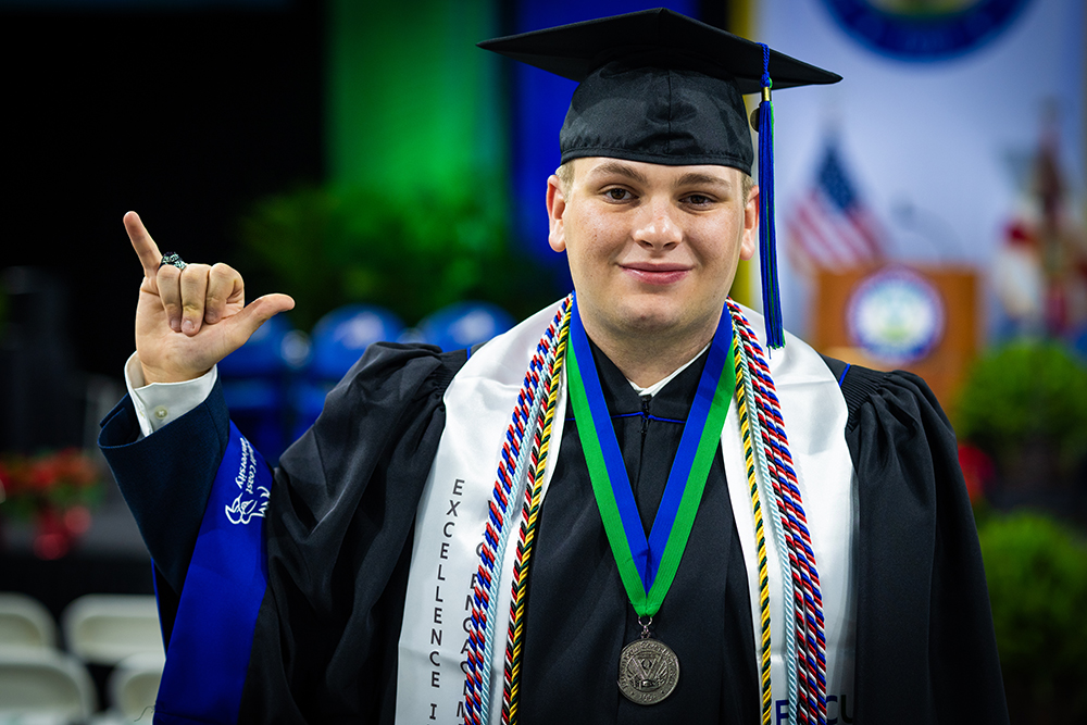 Man in commencement robe and sashes making the Wings Up hand gesture