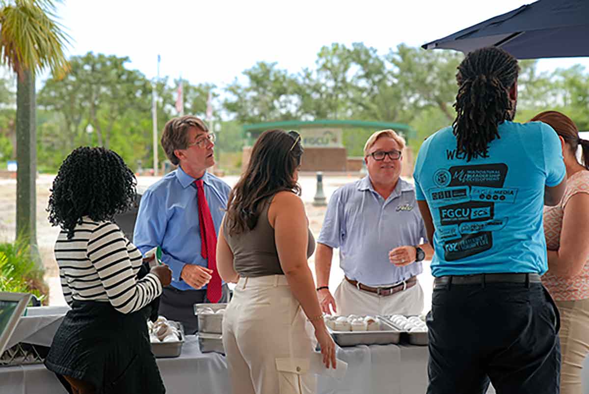 A man in a blue button-up shirt and red tie stands next to a man in a blue FGCU polo, serving ice cream to a line of students