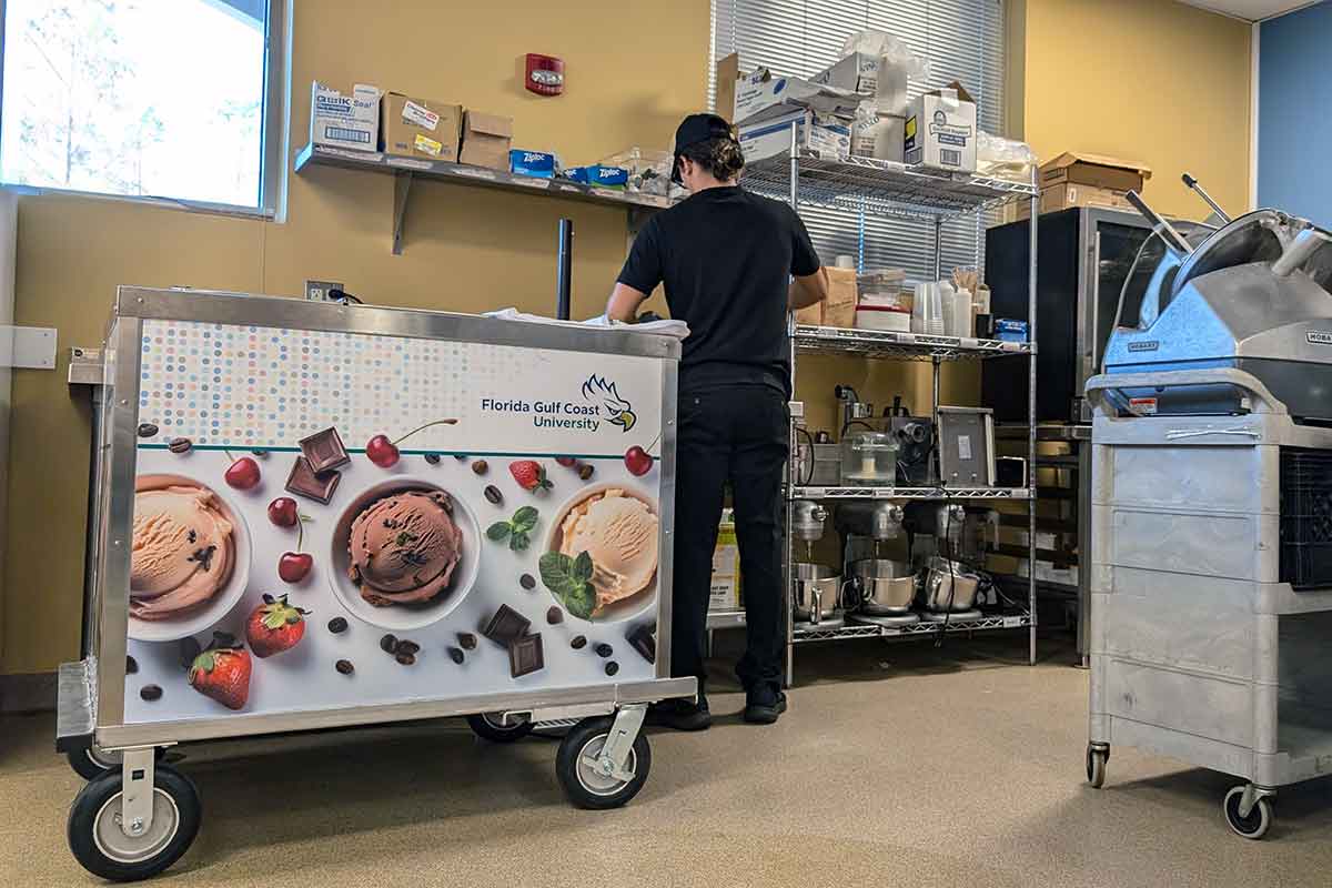 A branded ice cream cart on wheels in a kitchen, with a worker dressed in a black hat, black polo shirt and black pants faces away from the camera