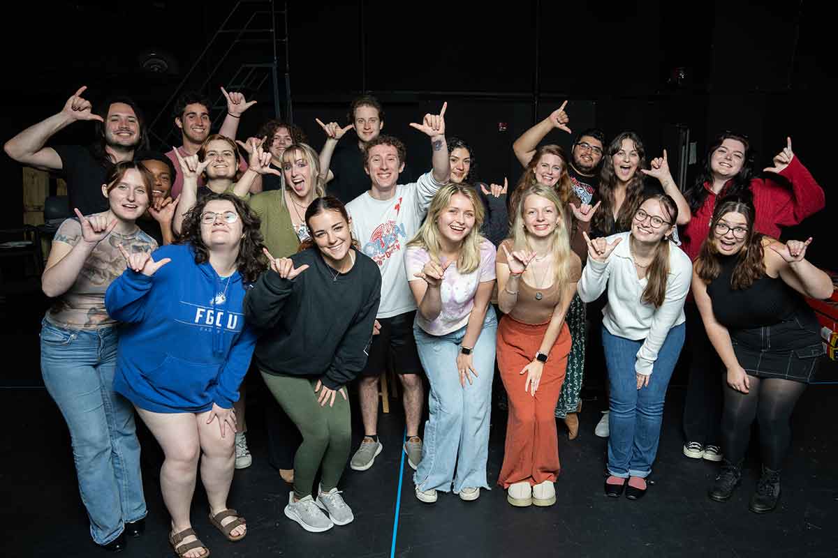 A large group photo in a black box theatre, with everyone holding up the FGCU Wings Up gesture