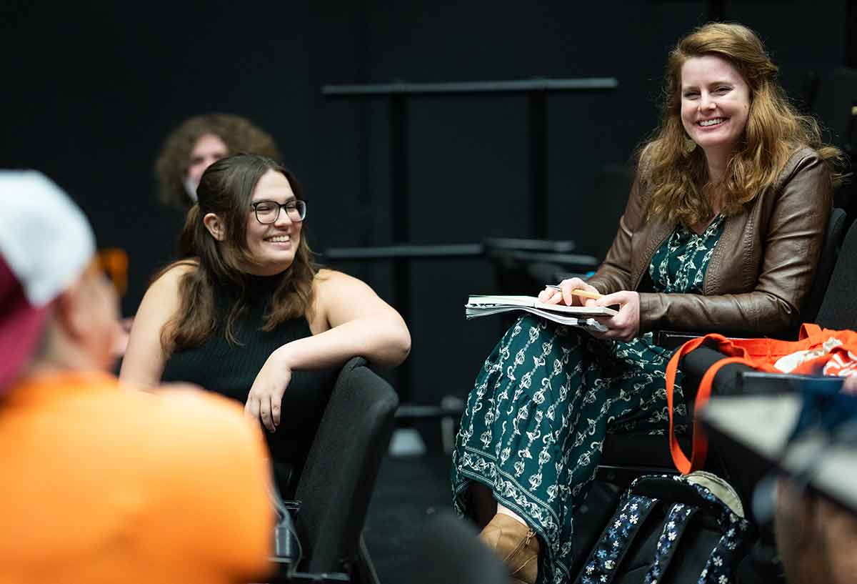 A woman in a green dress and brown leather jacket sits in a black box theater with a small group of students