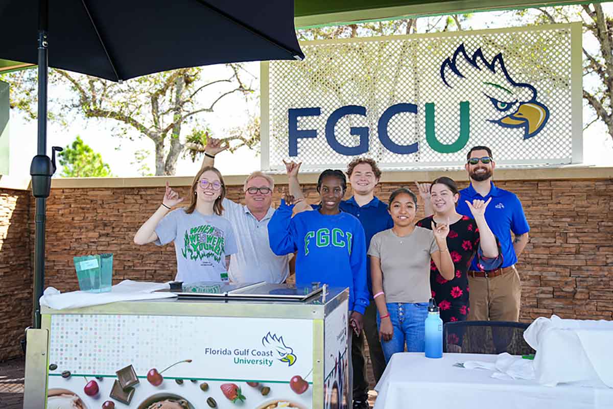 A group of staff and students make the FGCU Wings Up gesture while standing in front of the branded ice cream cart