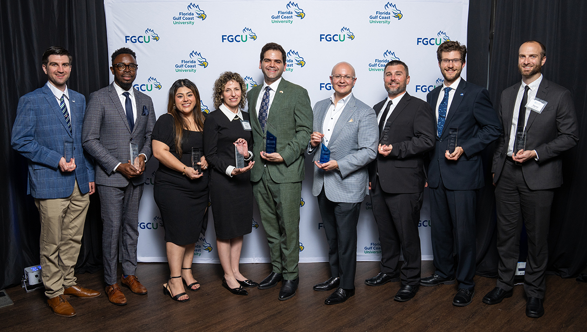 Seven men and two women standing in front of a step-and-repeat holding awards