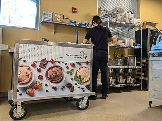 A branded ice cream cart on wheels in a kitchen, with a worker dressed in a black hat, black polo shirt and black pants faces away from the camera