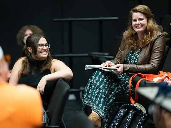 A woman in a green dress and brown leather jacket sits in a black box theater with a small group of students