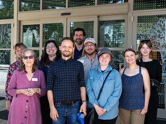 A group of nine people pose in front of an artistic window design of birds and trees and a grid of dots