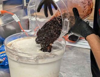 A bucket of cream sits on a metal table as gloved hands pour a bucket of fruit into the cream