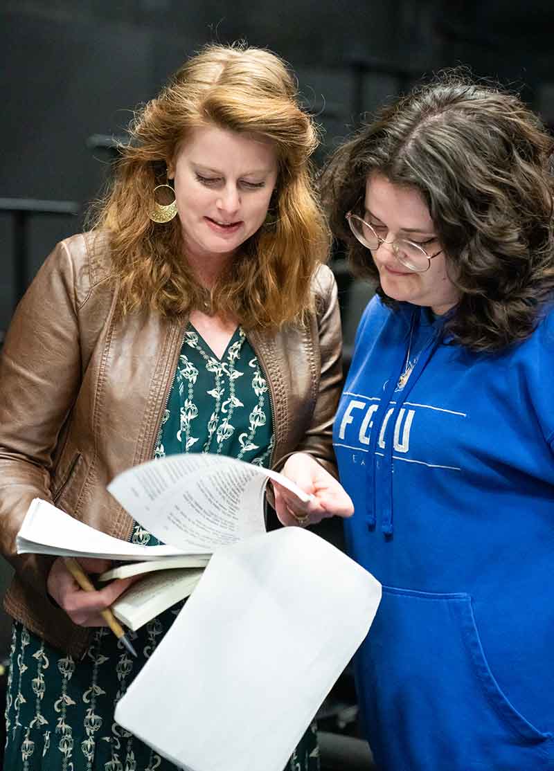 A woman in a green dress and brown leather jacket holds a script in her hand and shares it with a woman in a blue FGCU t-shirt