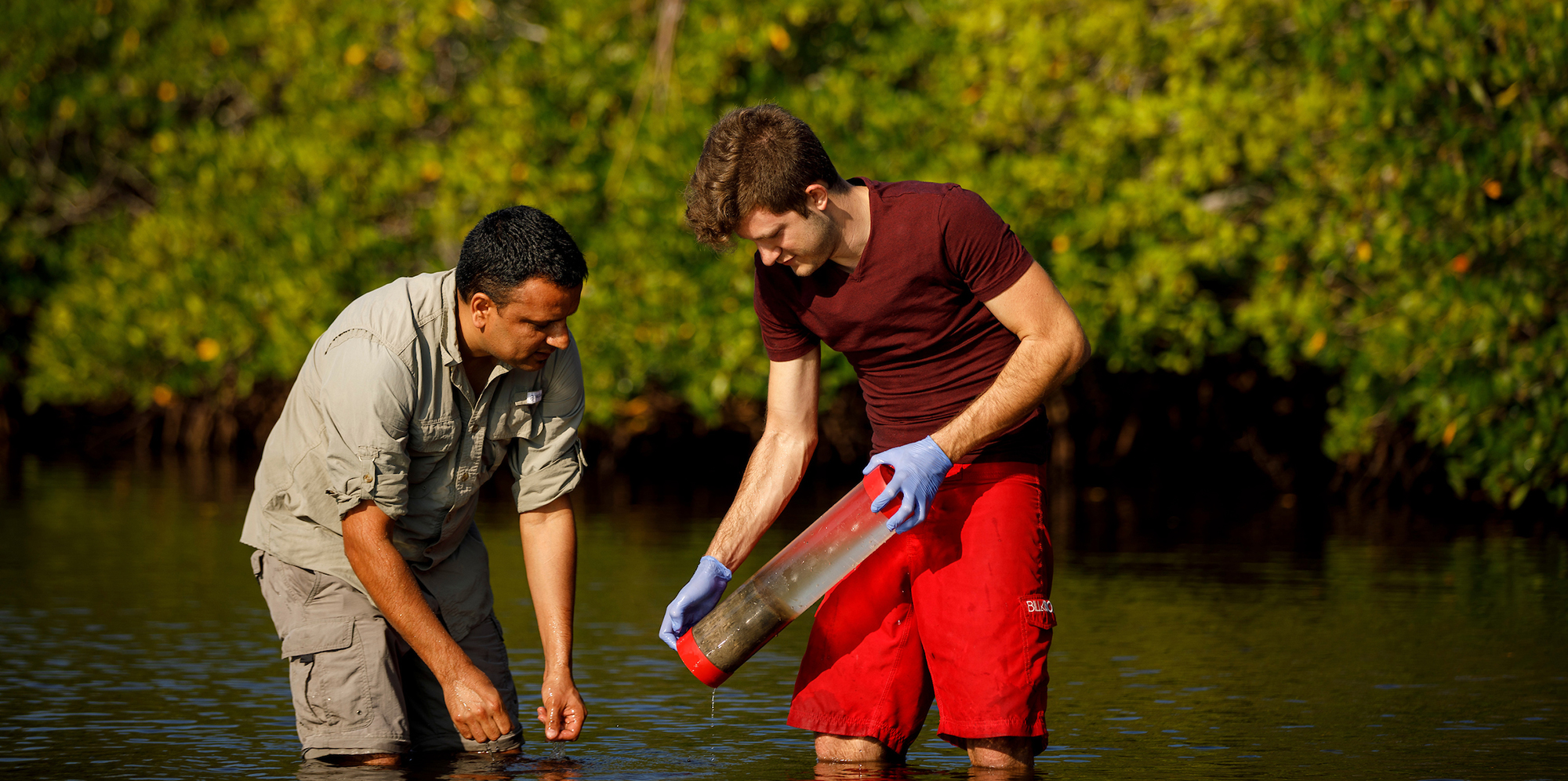 Two men standing in water looking at plastic tube of water and sediment