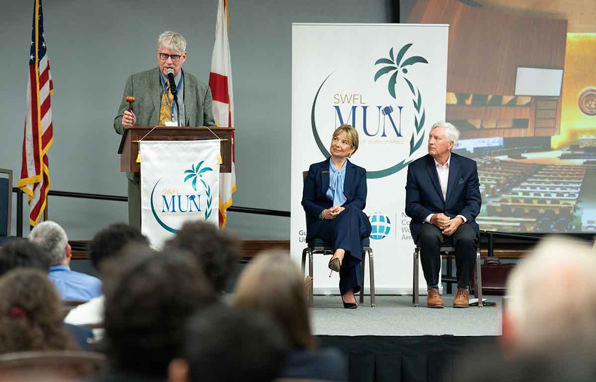 A white-haired man in a grey suit stands at a podium with a gavel and microphone as two seated people in business attire look on