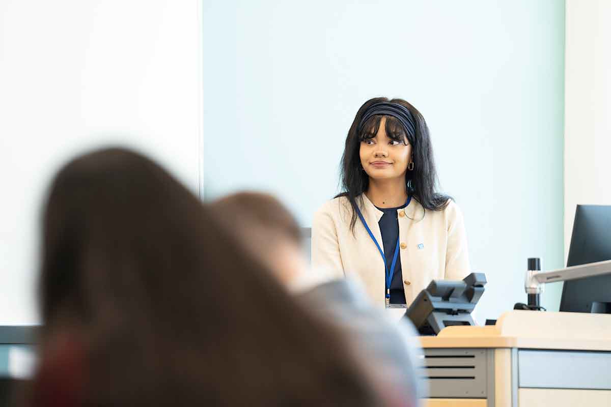 A woman with dark hair in a white blazer with black trim sits behind a classroom podium