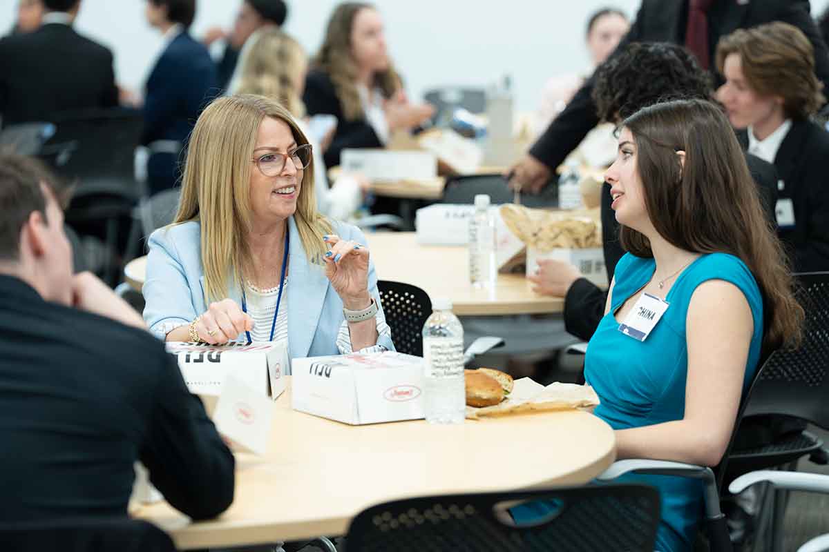 A blonde woman in a light blue blazer and glasses seated at a table talks to a young woman in a teal dress with a nametag that says China