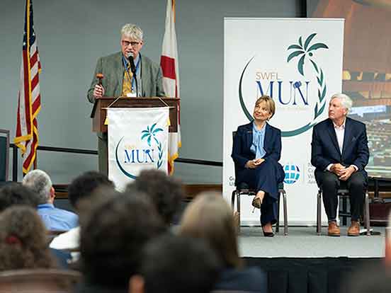 A white-haired man in a grey suit stands at a podium with a gavel and microphone as two seated people in business attire look on