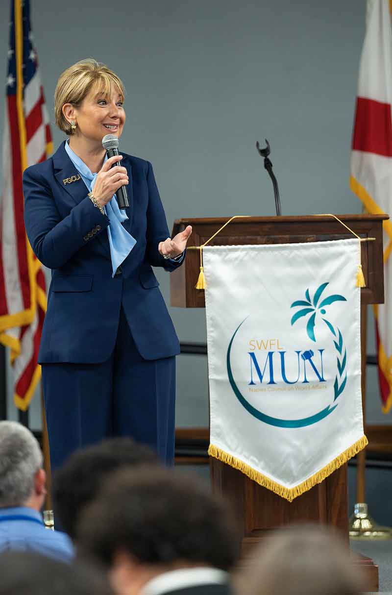 A woman in a navy blue pantsuit and baby blue shirt stands in front of a podium holding a microphone and speaking to a crowd