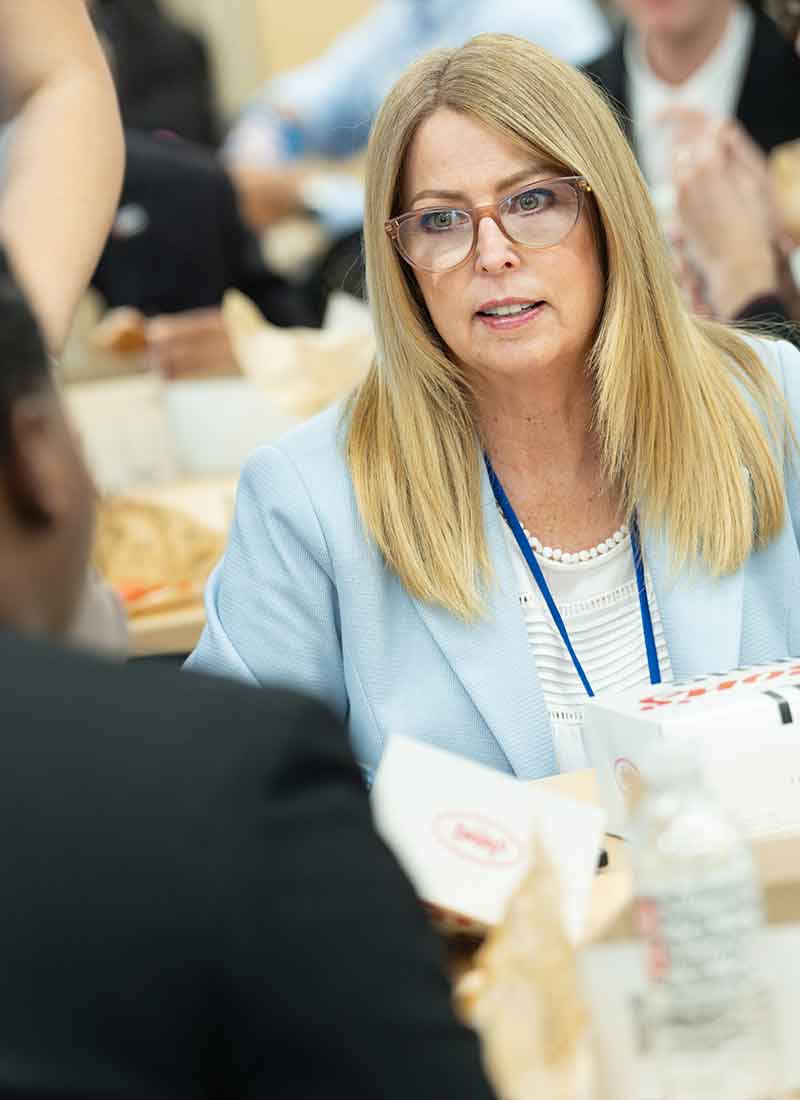 A blonde woman in a light blue blazer and glasses talks to someone out of the shot at a table